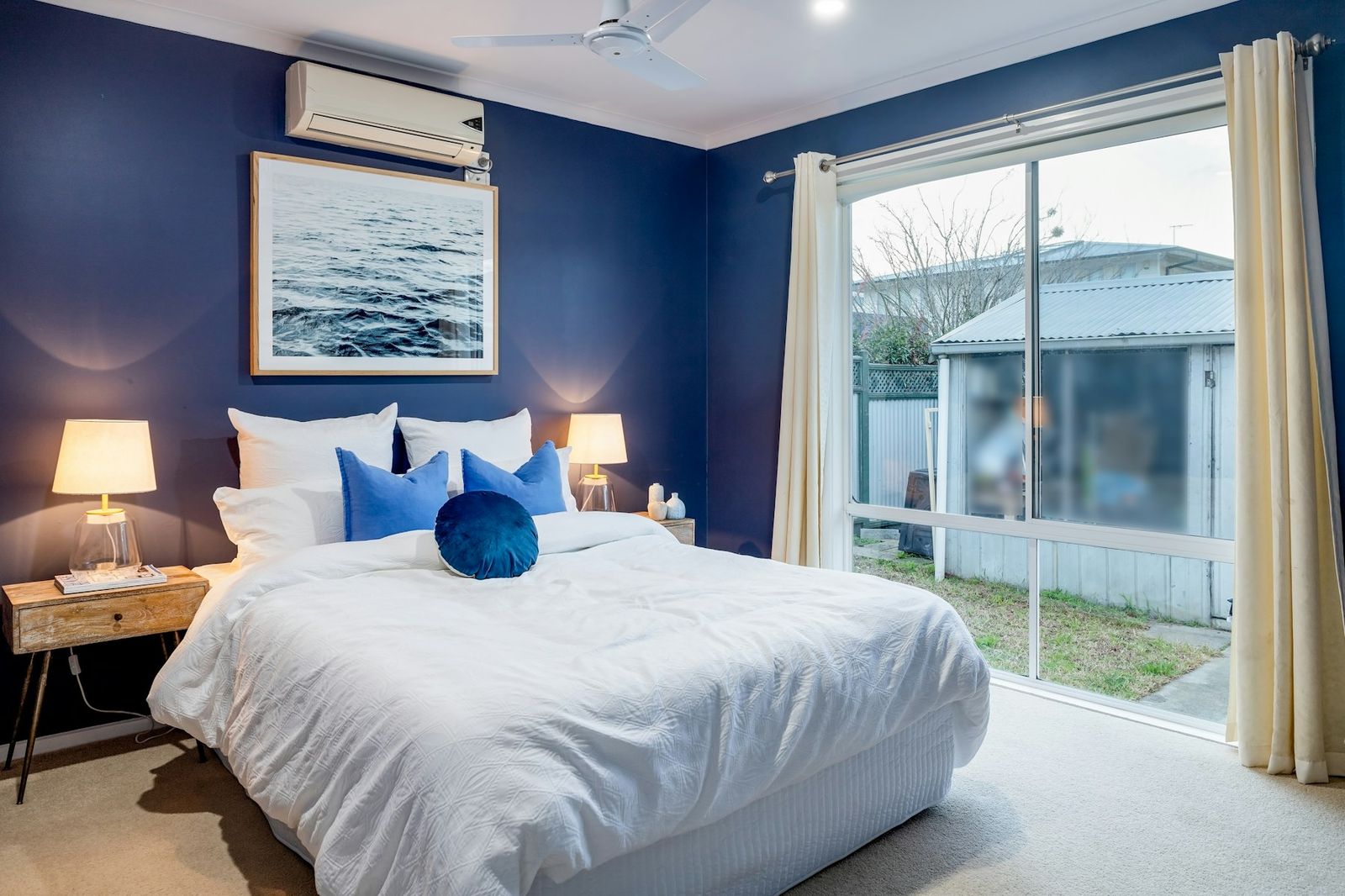 A bedroom with a wall-mounted air conditioning unit above the window and a garden beyond